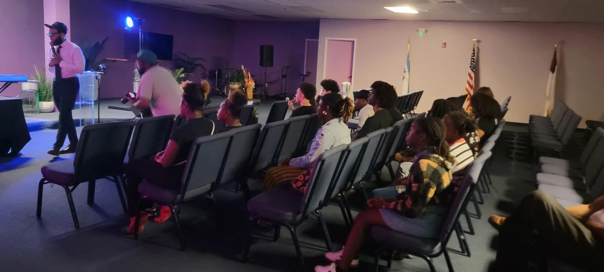 A group of people sit in rows of chairs facing forward in a dimly lit room, while a few stand near the front; flags are visible in the background.
