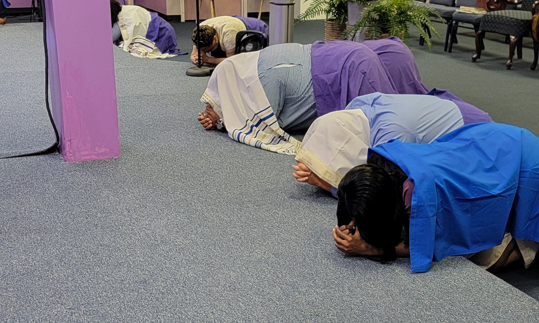 Four people in robes kneel and bow on a carpeted floor in a church or worship setting near a purple column.