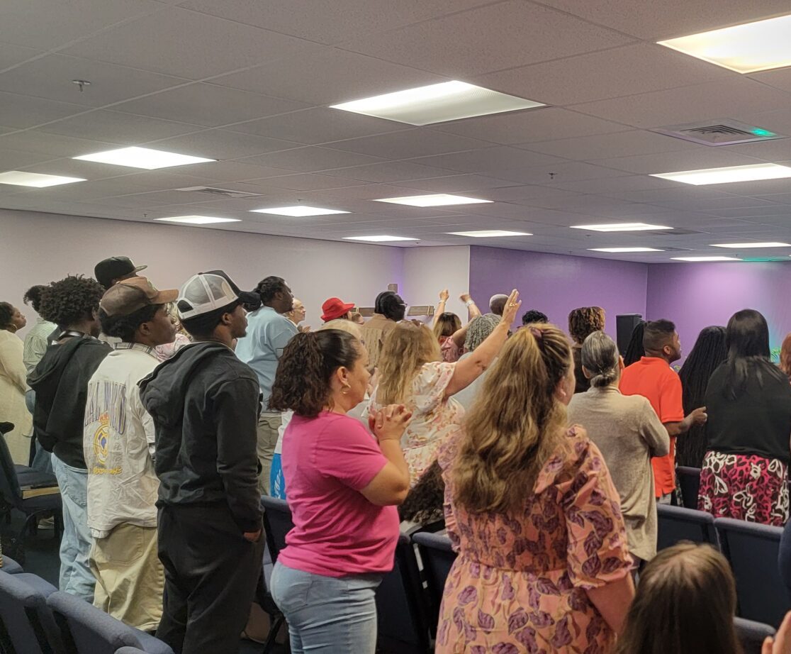A group of people stand in rows of chairs facing a screen with a cross image in a room with purple walls and overhead lights.